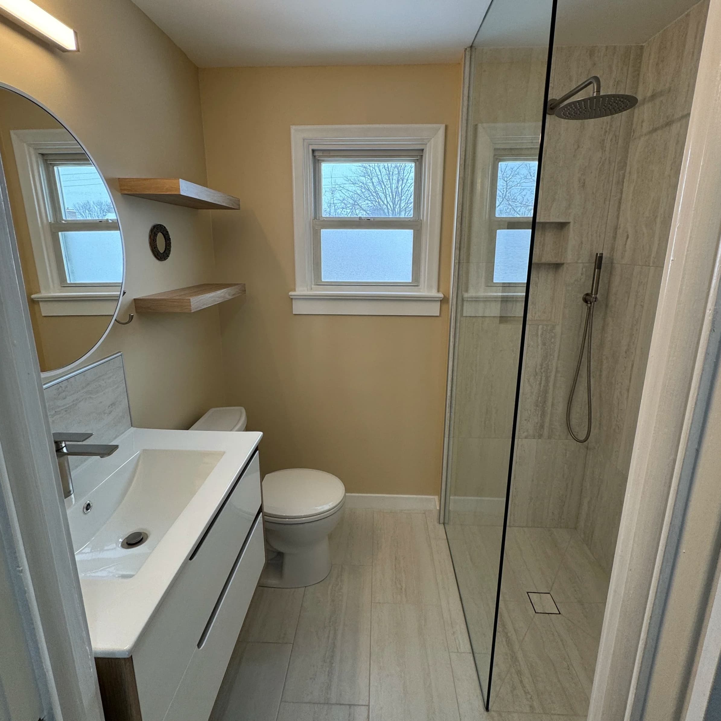 Small bathroom with floating white vanity, floating wood shelves, and walk-in tile shower.