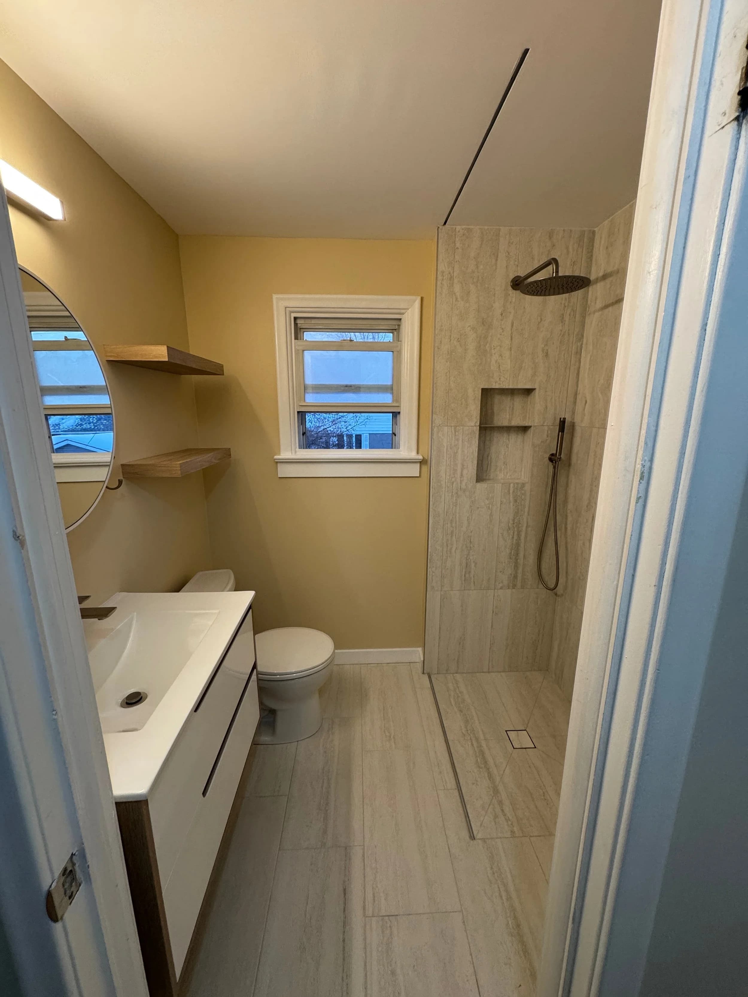 Compact bathroom with floating white vanity, round mirror, and walk-in tile shower lit by a large window.
